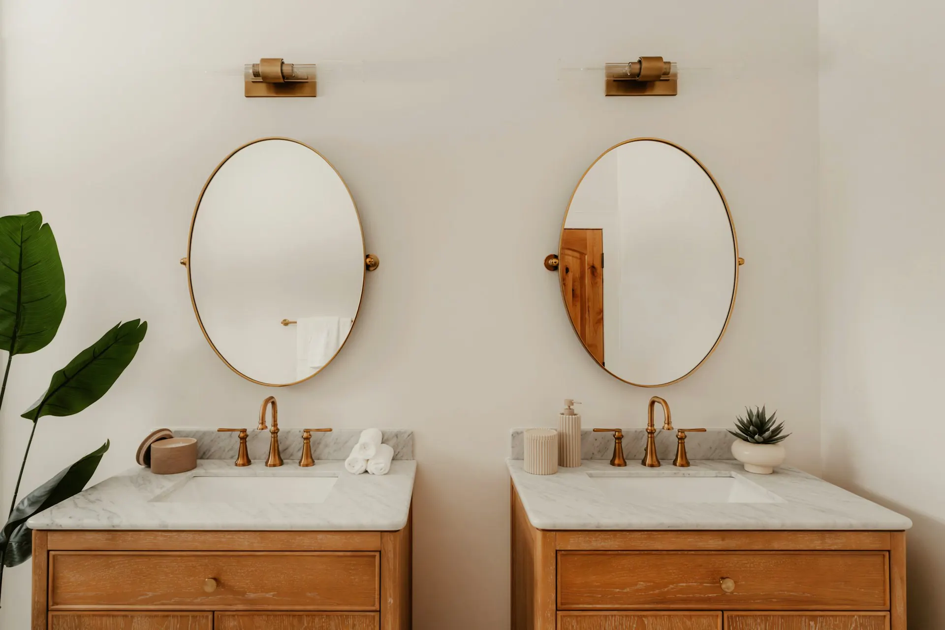 Two wooden bathroom vanities with oval mirrors and gold fixtures — double vanity primary bath